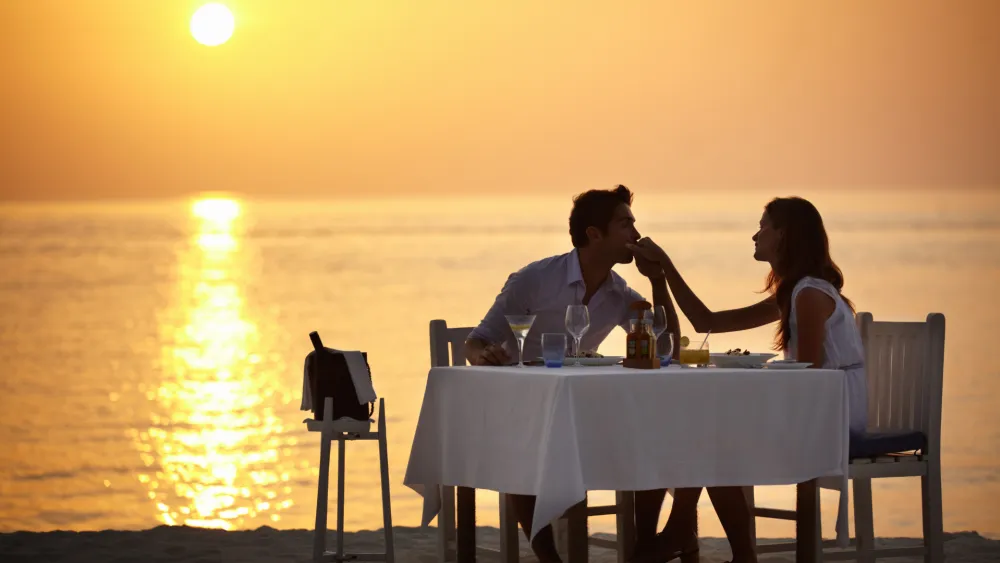 A couple sharing a romantic sunset dinner on the beach, toasting with wine as waves glow in the golden hour.