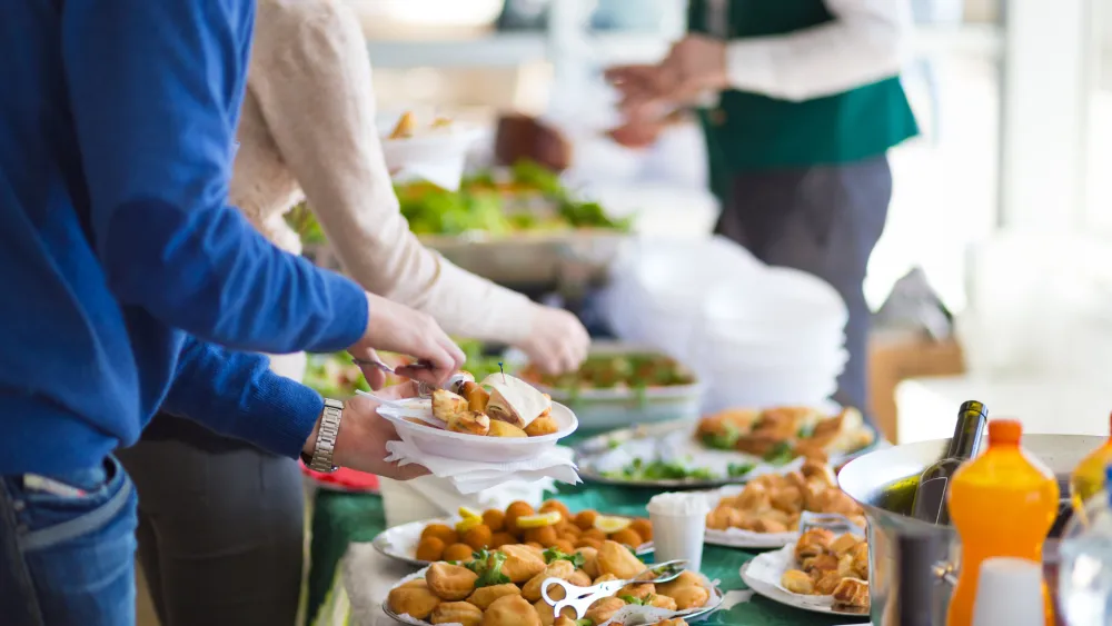 A group of people serving food at a communal buffet line with various dishes laid out on a table.