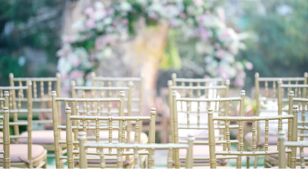 Golden chiavari chairs set up outdoors at a wedding-style event, with colorful floral decor blurred in the background.