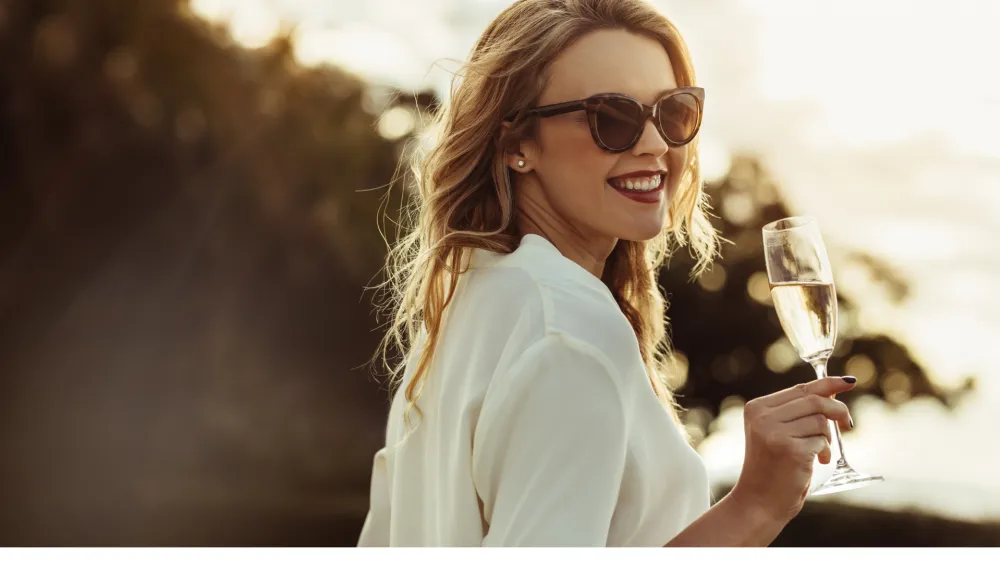 A woman in sunglasses smiles while holding a champagne flute outdoors at sunset, wearing a white blouse.
