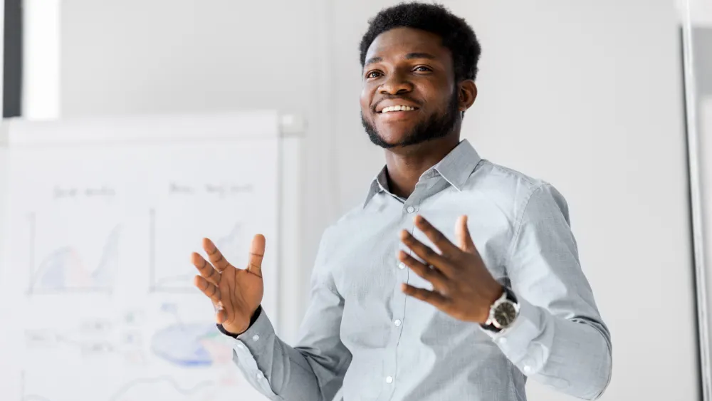 A man presents and gestures in front of a chart, explaining data on a whiteboard.