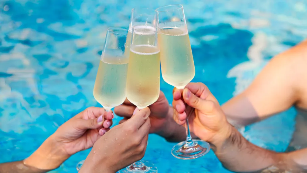 Four people clink sparkling champagne glasses by a sunny pool, celebrating together with bubbly drinks.