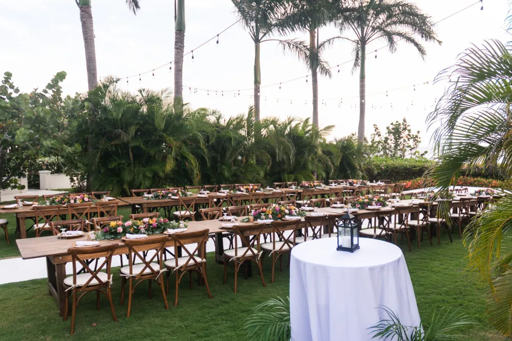 An outdoor garden banquet setup with long wooden tables, colorful flowers, lanterns, string lights, and tall palm trees.
