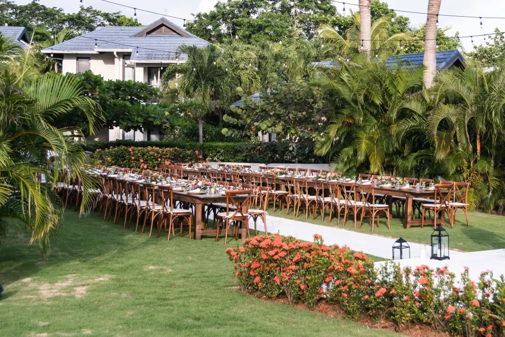 Tropical garden wedding setup with long tables, wooden chairs, floral centerpieces, string lights, and a villa backdrop.