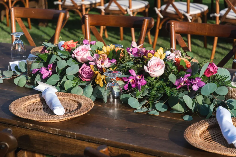 A rustic outdoor table set with a lush centerpiece of pink, purple, and yellow flowers, eucalyptus, and rolled napkins.