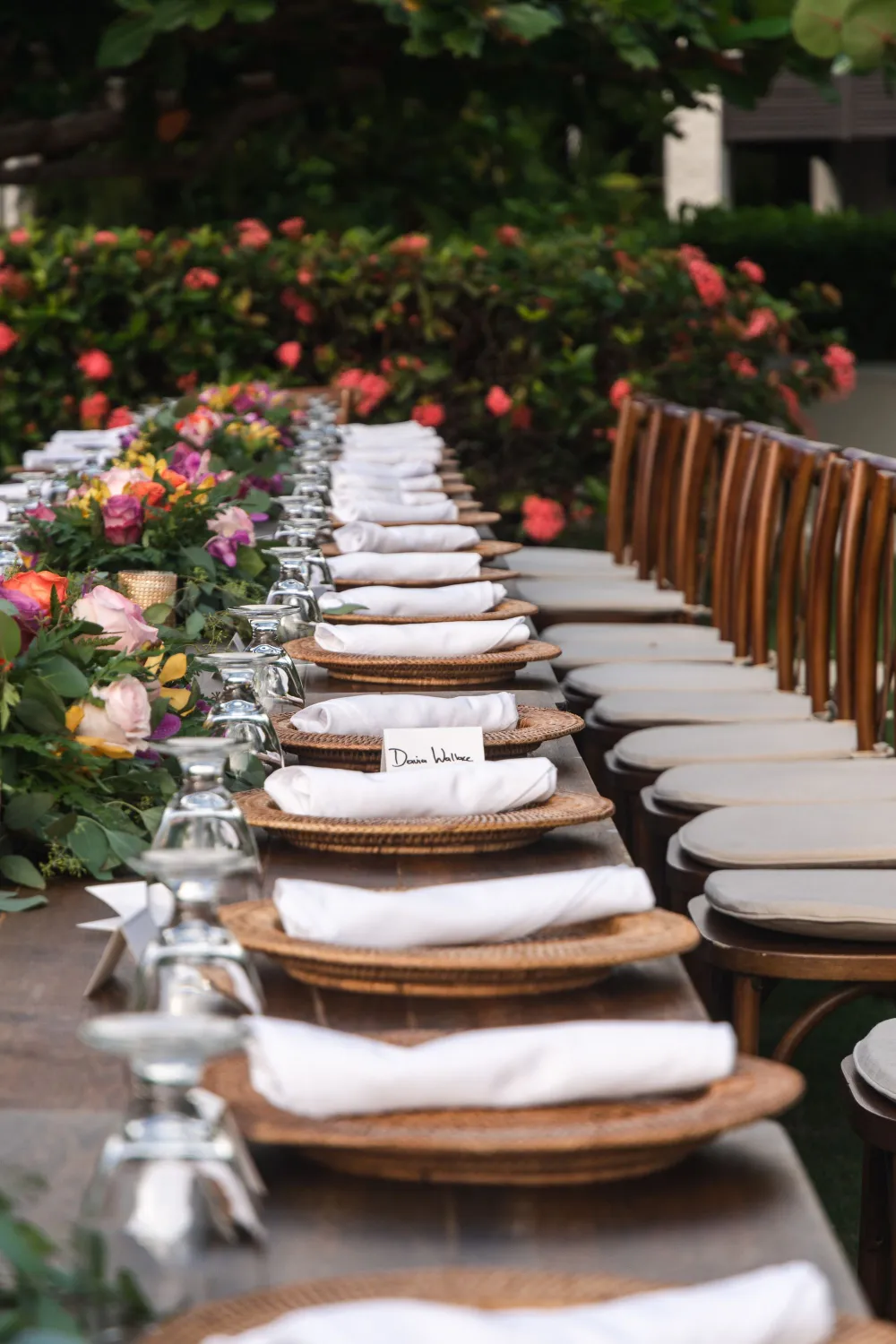 A long outdoor table setup with white plates, napkins, and glassware ready for a meal, surrounded by flower arrangements and greenery at a garden party.