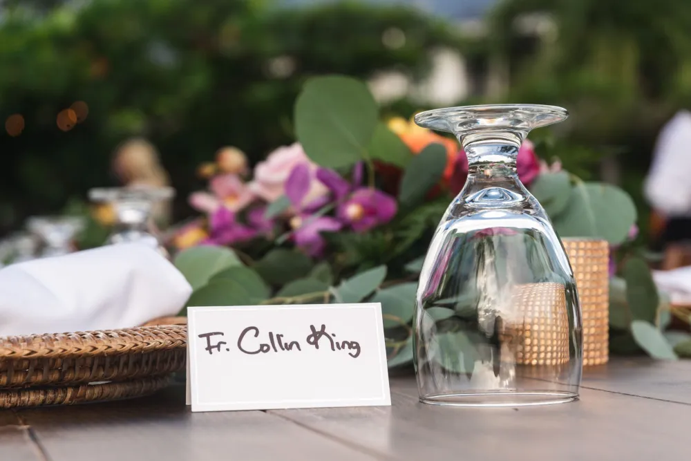 A table set for a formal event with a place card labeled “Fr. Colin King,” a glass upside down, floral centerpieces, and elegant decor.