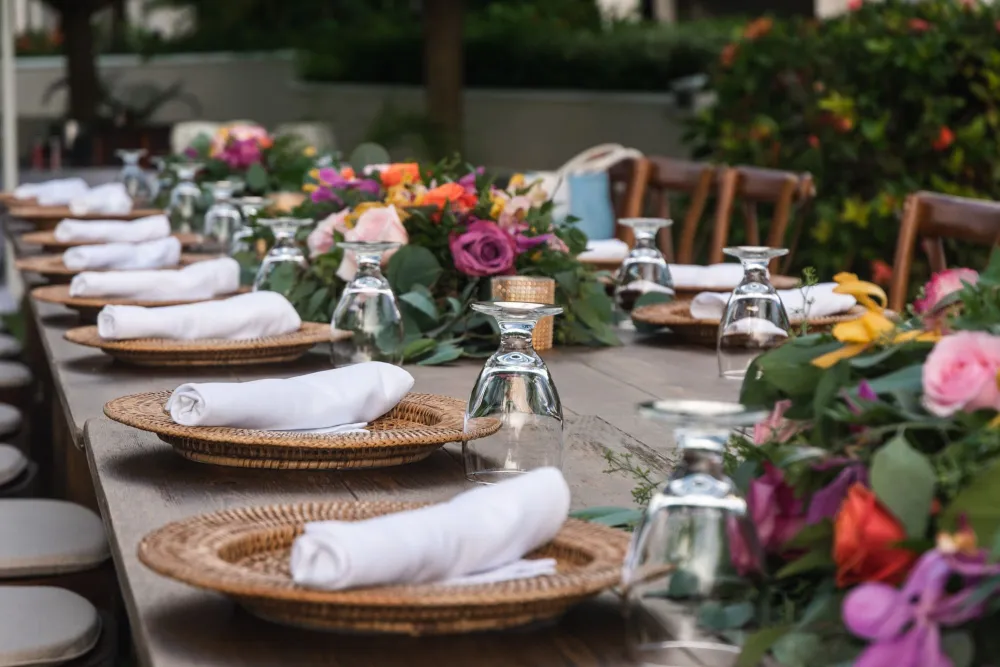 An outdoor dining table set with glassware, folded napkins, and colorful flower arrangements in the center.