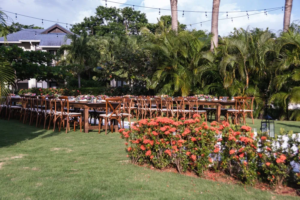 An outdoor garden banquet setup with a long table, wooden chairs, bright flowers, palm trees, and string lights overhead.