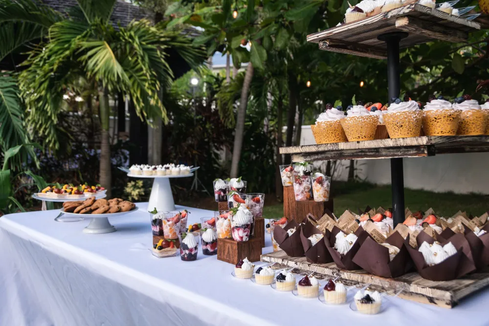 Outdoor dessert table with cupcakes and fruit parfait cups topped with cream, set in a lush garden among palm leaves.