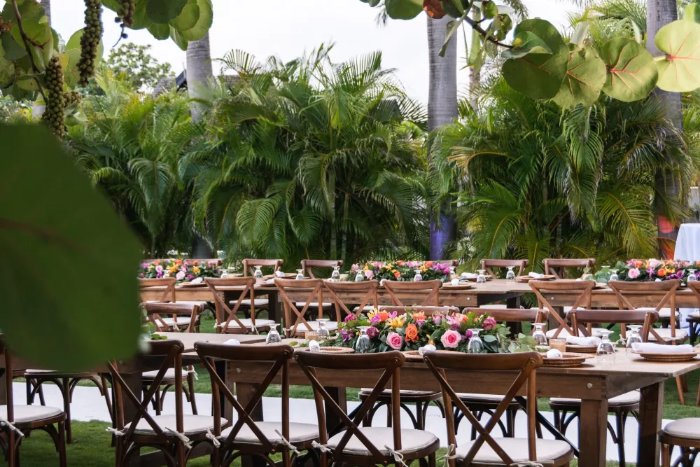 An outdoor tropical dining setup with wooden chairs, long tables, colorful flower centerpieces, and lush palm trees behind them.