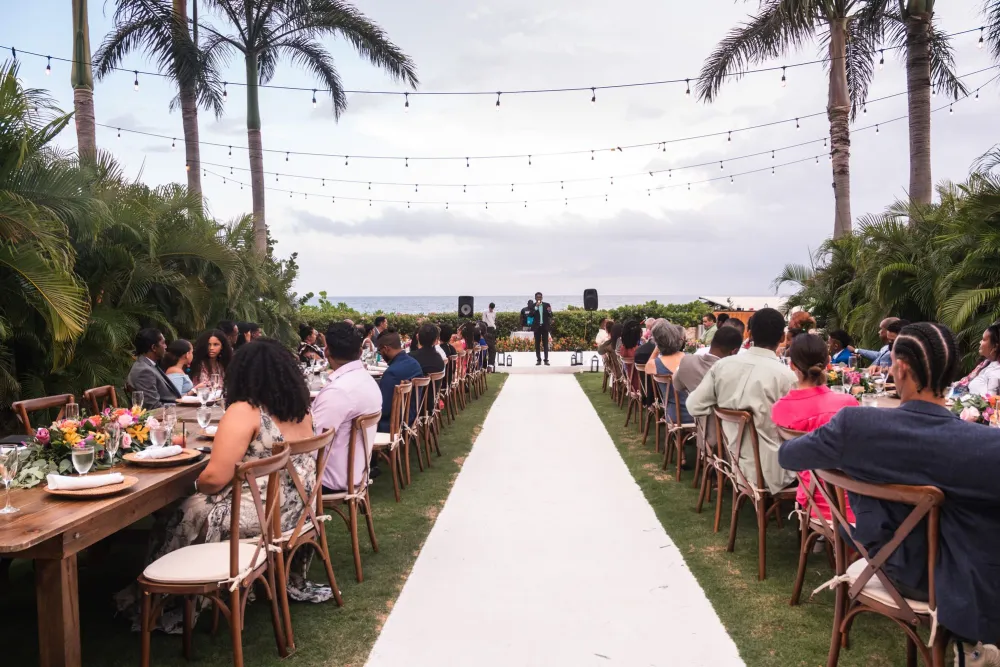 Outdoor beach wedding reception with guests seated on both sides of a garden aisle beneath palm trees and string lights.