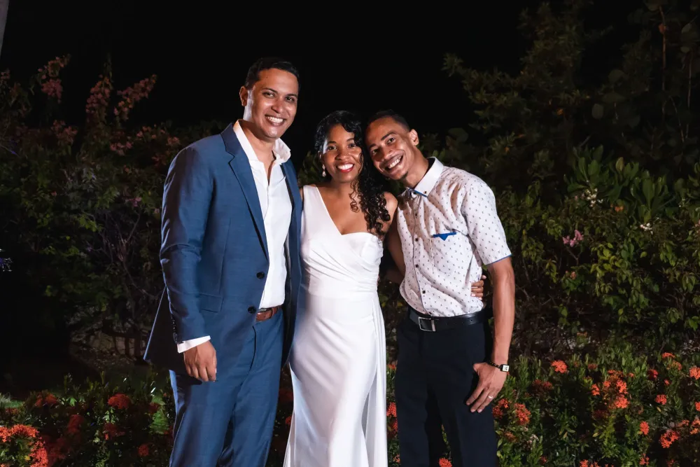 A smiling trio in formalwear posing outdoors at night among flowers and trees.