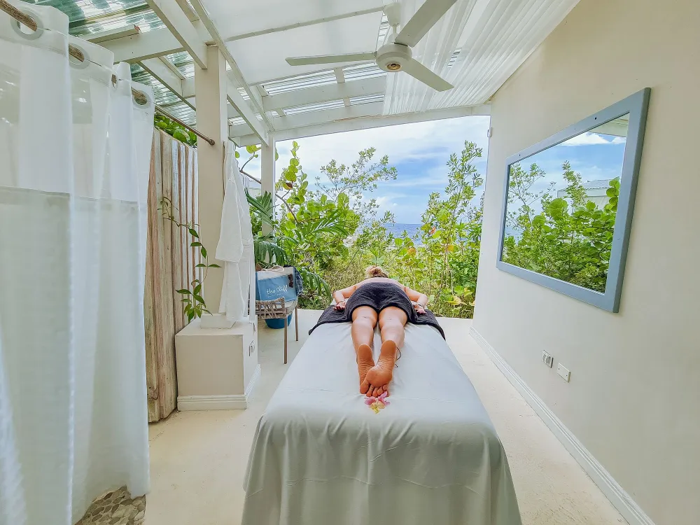 A person is lying face down on a massage table in a bright, airy spa room with a view of greenery and ocean beyond, relaxing.