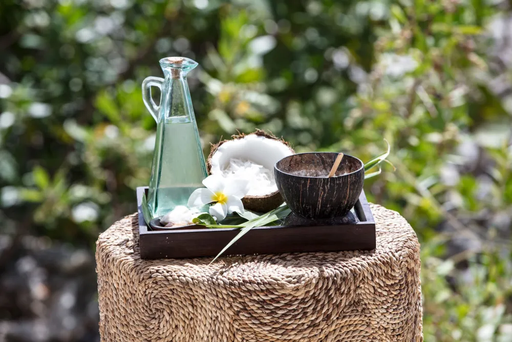 A glass bottle with a stopper, a small wooden bowl, a coconut half, a wooden tray, a flower, and a few glass cups on a woven stool outdoors, serene.