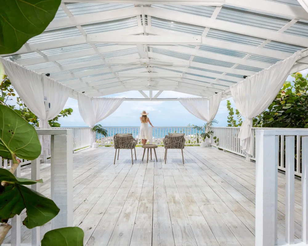 A person stands on a bright, white wooden deck under a white canopy, facing the ocean with chairs arranged around a table—tropical and serene.
