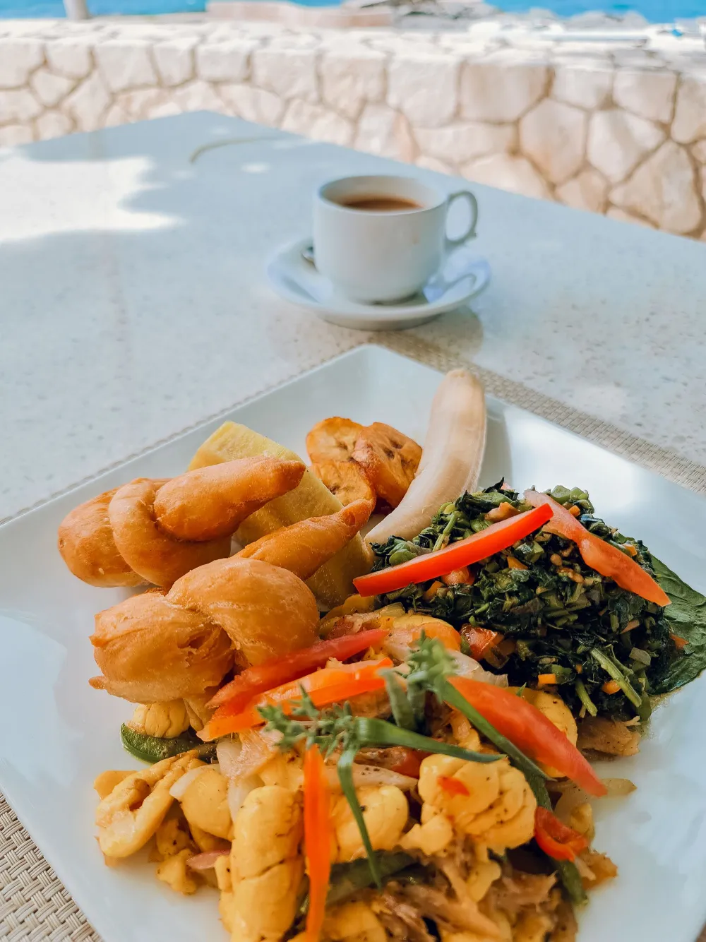 Delicious plate with croissants, sautéed vegetables, greens, and chicken, plus a cup of coffee in the background, on a sunny outdoor table.