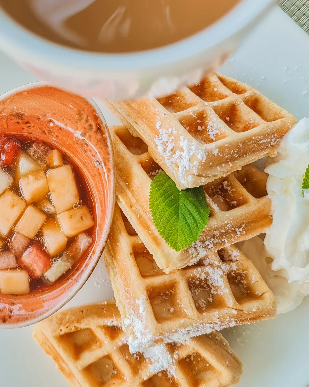 Delicious waffles stacked with powdered sugar, maple drizzle, cubed fruits in a side dip, and a dollop of whipped cream with a mint leaf, yum.