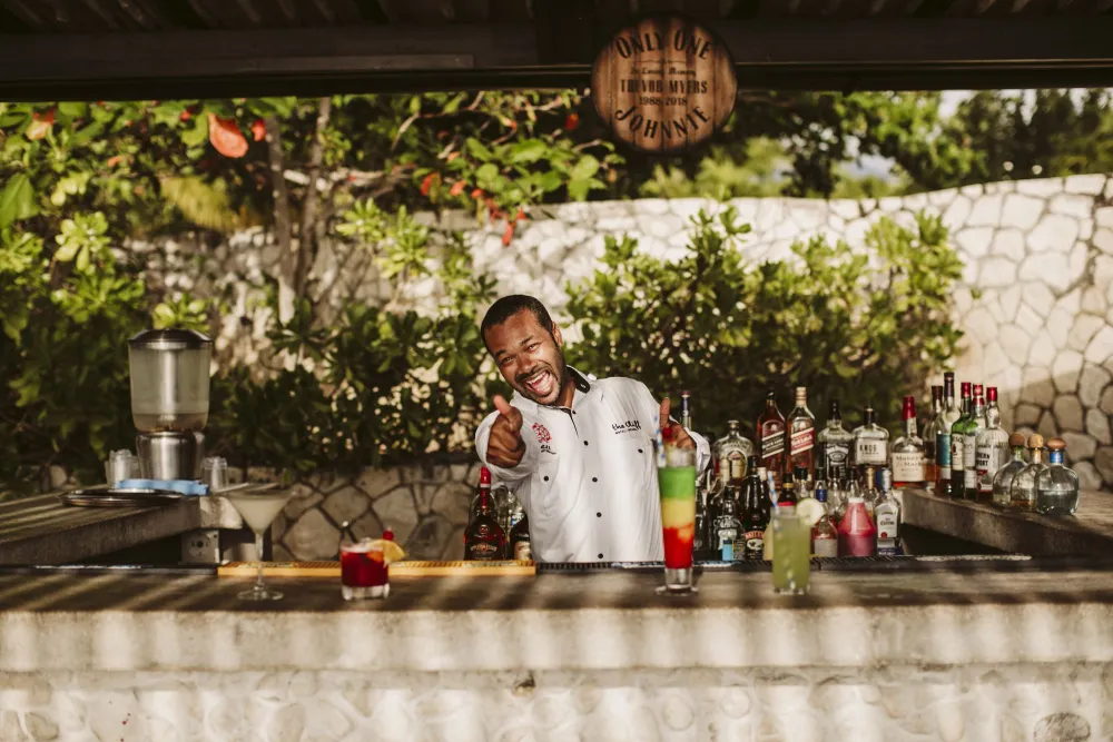 A smiling bartender in a white shirt behind a tropical outdoor bar, pointing at the camera, with colorful bottles and drinks on the counter.
