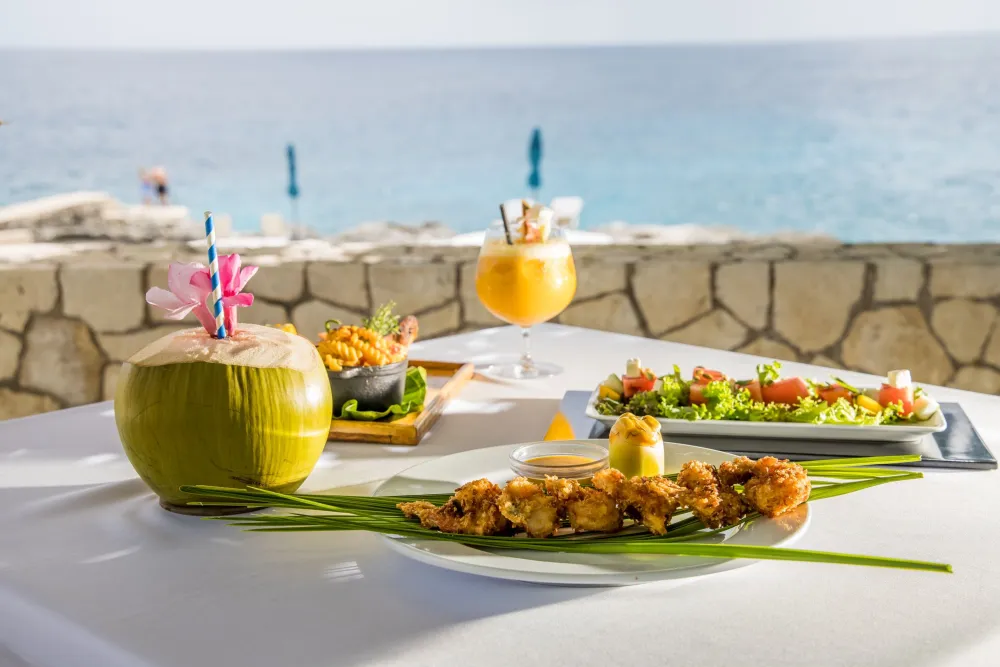 A sunny seaside meal with tropical drinks, grilled skewers, and fresh salads on a white-tabletop overlooking the ocean.