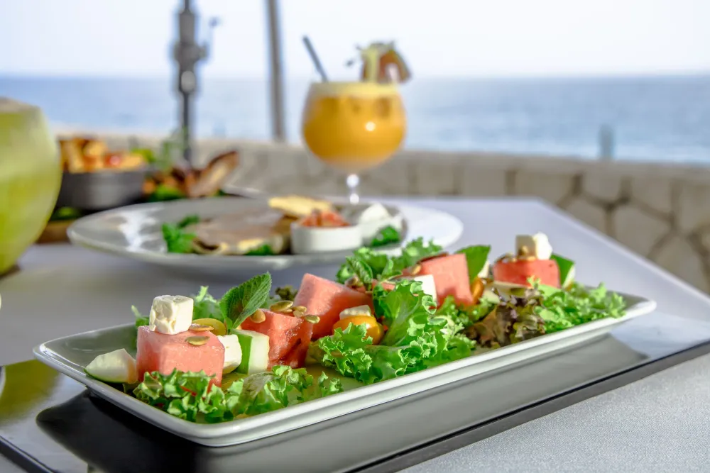 A vibrant plate of salad with watermelon and feta chunks on lettuce, garnished with herbs, on a seaside table; a drink and more food in the background.