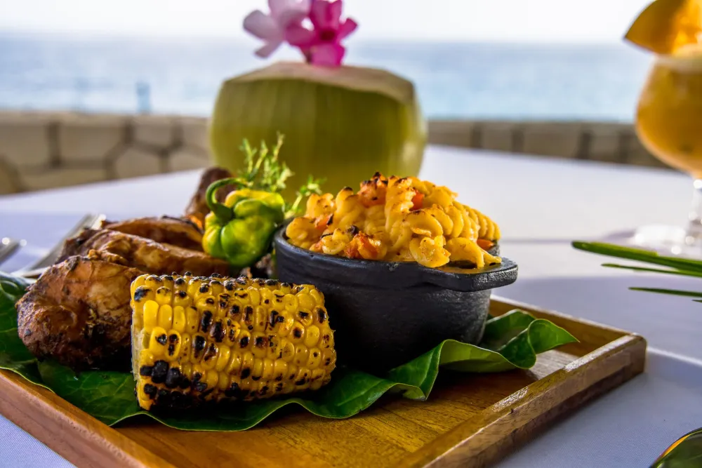 Tropical beachside meal: grilled meat, corn on the cob, two bowls of golden rice with herbs, all served on a wooden board with a coconut drink.