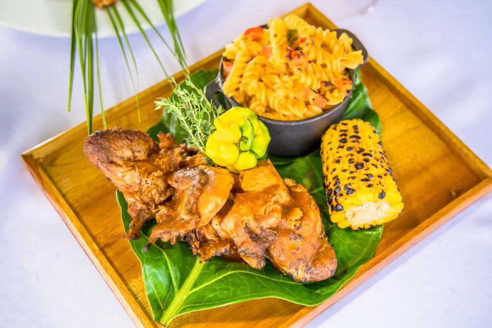 A plated meal featuring fried chicken pieces on a banana leaf, a side of corn with black sesame seeds, and a bowl of fried rice on a wooden tray.