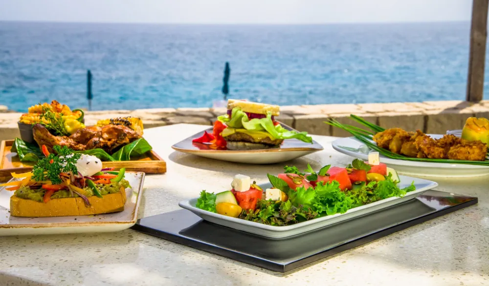 An oceanfront spread of colorful dishes on a white table: grilled sandwiches, fresh salads, and greens, with a blue sea backdrop.