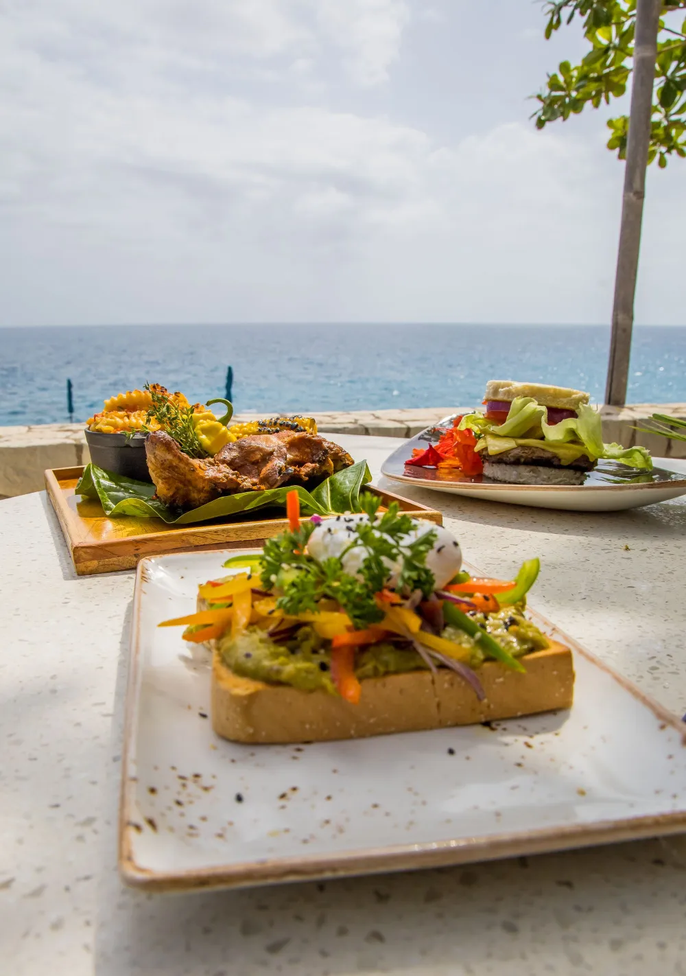 A seaside meal with three plated dishes on a light table, featuring toasted bread piles topped with veggies and herbs, and a bright ocean backdrop. (Note: ending punctuation should be period.)