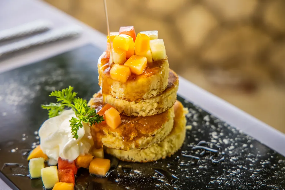 A stack of golden crumbed fried rounds topped with diced mango/papaya cubes, a dollop of cream, and a sprig of herbs, drizzled with syrup.