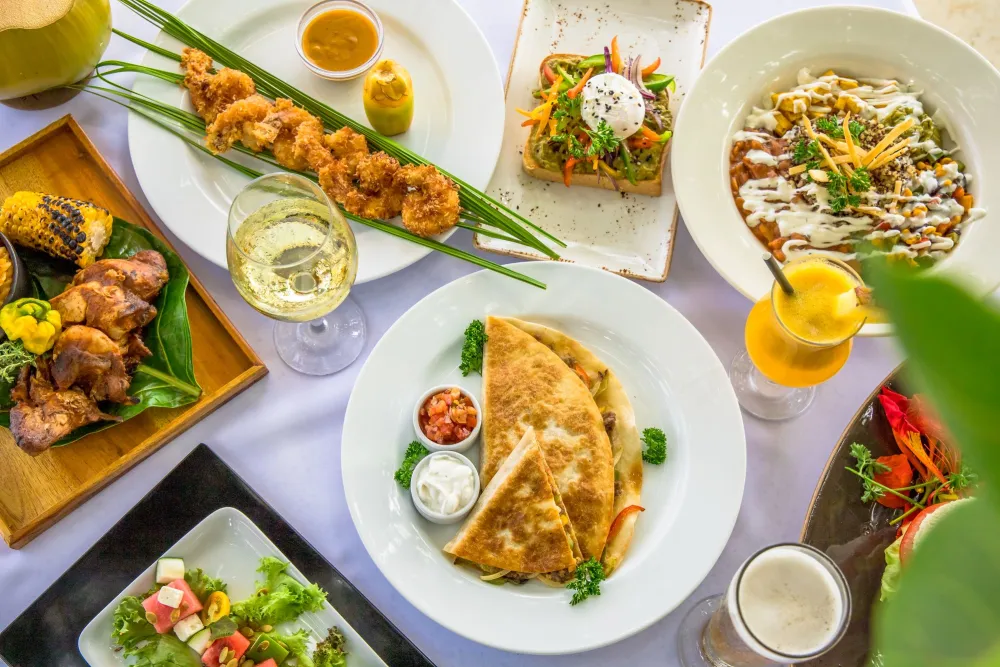 A colorful spread of foods—quesadillas, crispy skewers, veggie toast, salad bowls, dips and drinks—served on white plates.