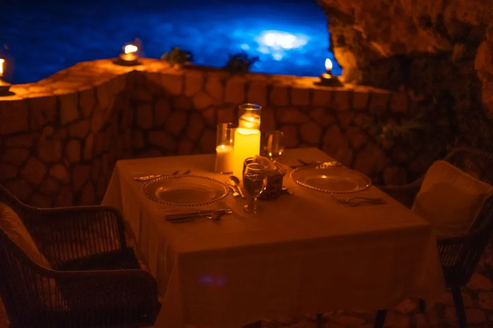 A candlelit romantic table setup on a terrace with a stone wall, dim lighting, two chairs, and a moonlit blue night sky.