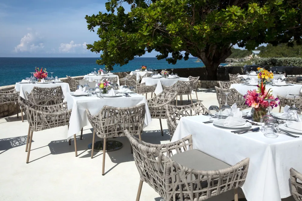 A beachside restaurant with white-tablecloth tables, wicker chairs, colorful flower centerpieces, and a view of the blue ocean under a leafy tree.