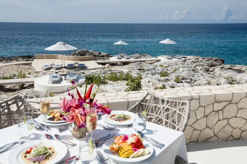 A seaside dining table set with white linens, flowers, and plates of food, stone terrace, sun umbrellas, and a blue ocean beyond the rocks. ending.