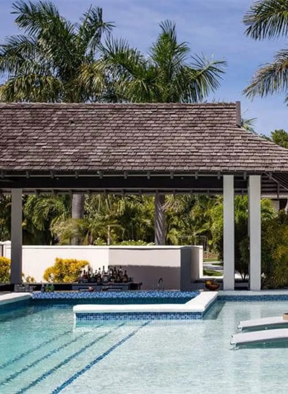 A resort-style pool with steps, a shaded gazebo, palm trees, and a sunny, tropical backdrop.