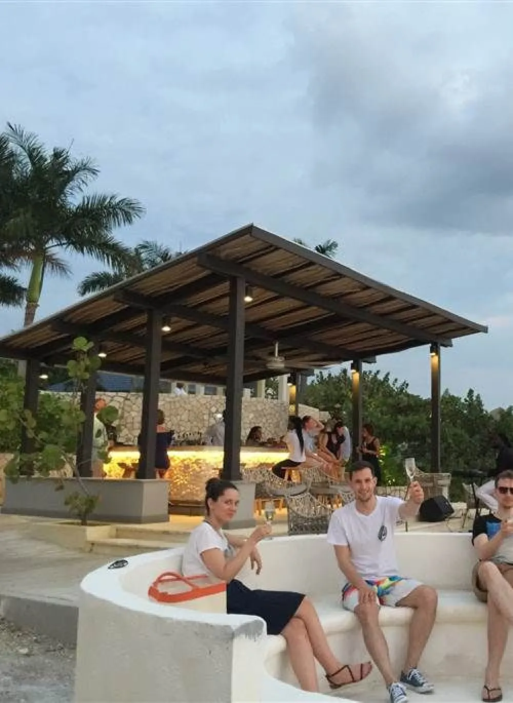 A few people sit and relax by a poolside bar with a wooden pavilion, palm trees, and a cloudy sky in the background.
