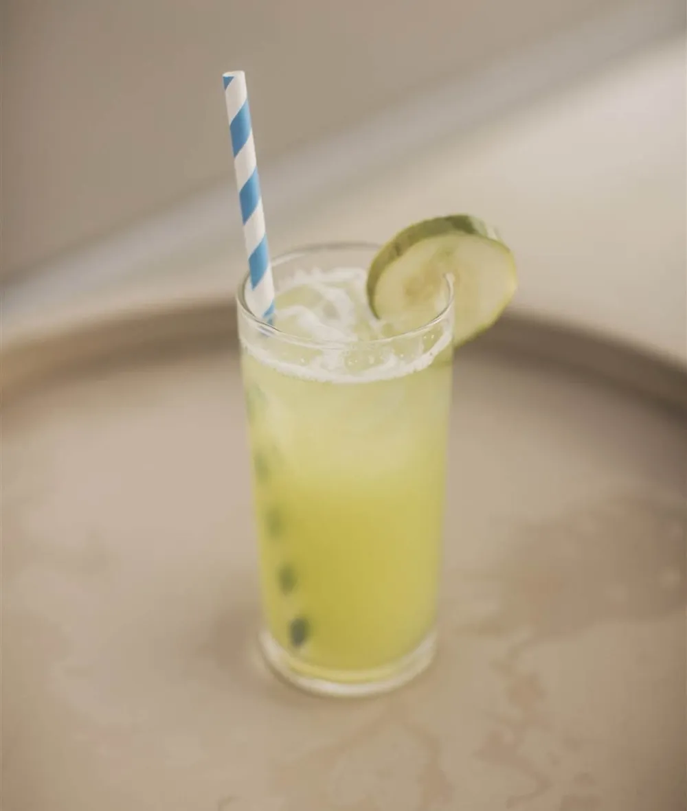 A tall glass of yellow lime drink with a striped blue straw and a lime wedge garnish, on a metal tray, looks refreshing.
