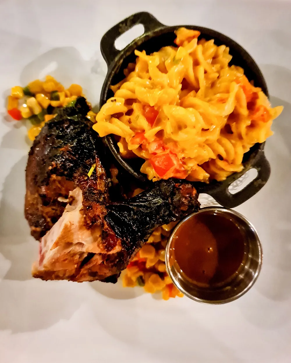 Smoked rib with charred crust, a side of cheesy macaroni, and a pool of dark barbecue sauce, plated over a bed of colorful corn and peppers.
