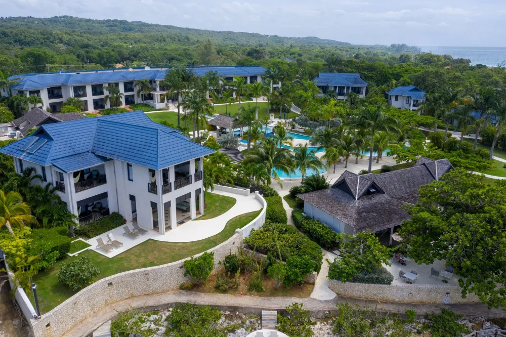 A resort with blue-roofed buildings, lush green gardens, a winding path, and a central pool area by the coast, tropical and serene.