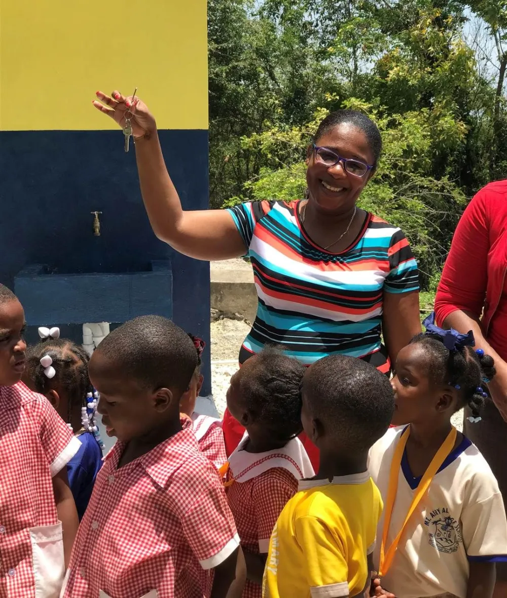 A smiling woman among children outside a colorful building, guiding them in a sunny outdoor setting, with everyone dressed in bright clothes.