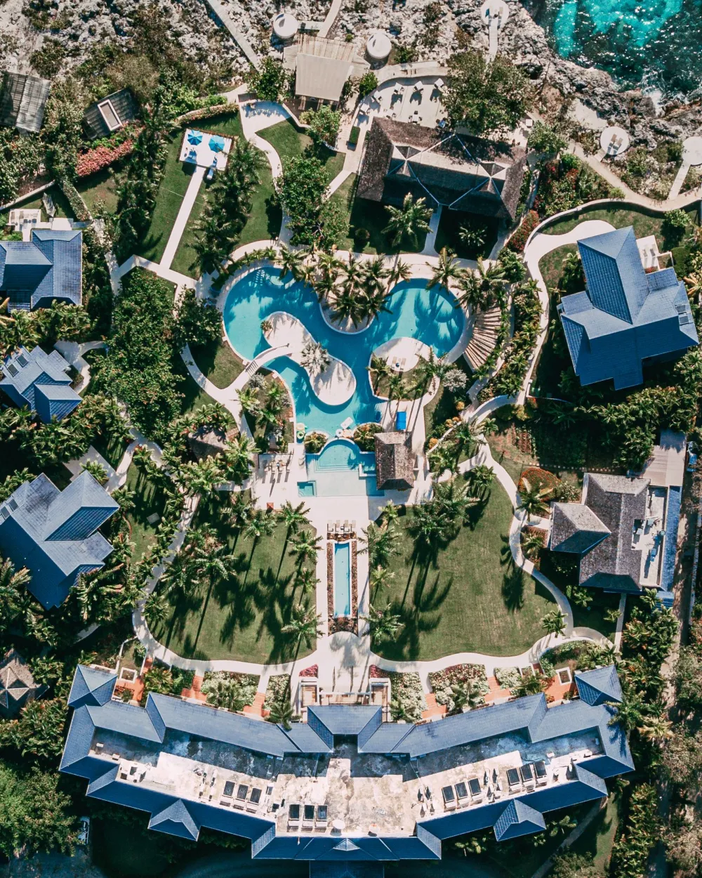 Aerial view of a resort with winding pools, palm trees, and surrounding villas, forming a heart/flower-like layout.