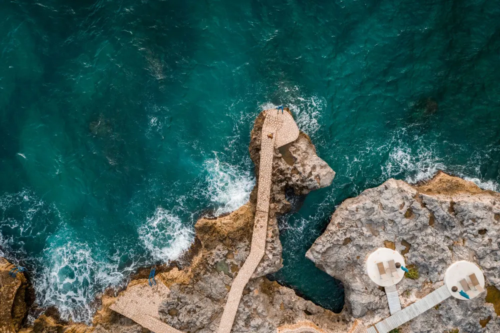 Aerial view of rugged rocky coastline with a concrete jetty extending into turquoise water, small white circular platforms at the edge, waves crashing below.
