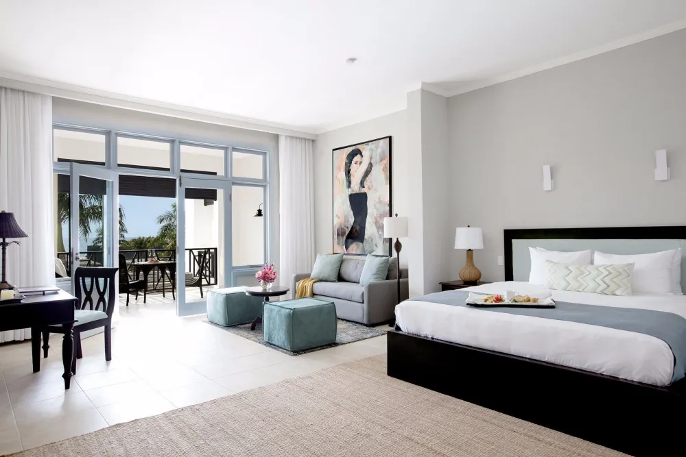 A bright hotel suite with a large bed, gray accent wall, a seating area, and glass doors opening to a balcony with outdoor views.