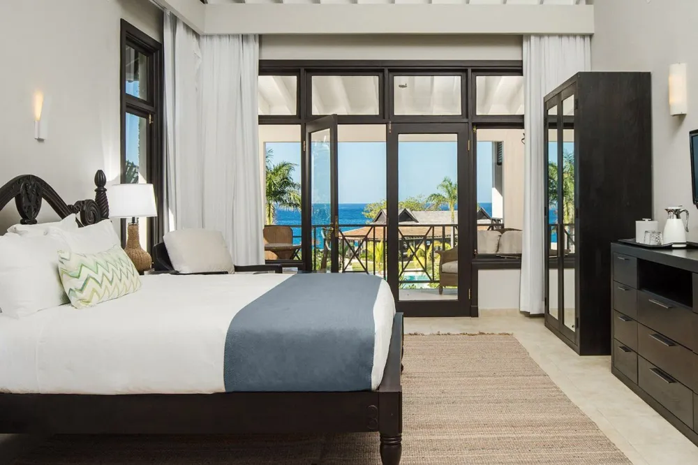 A hotel room with a dark wooden bed, white and blue bedding, a rug, and a balcony view of palm trees and the ocean through glass doors. Ending with a period.