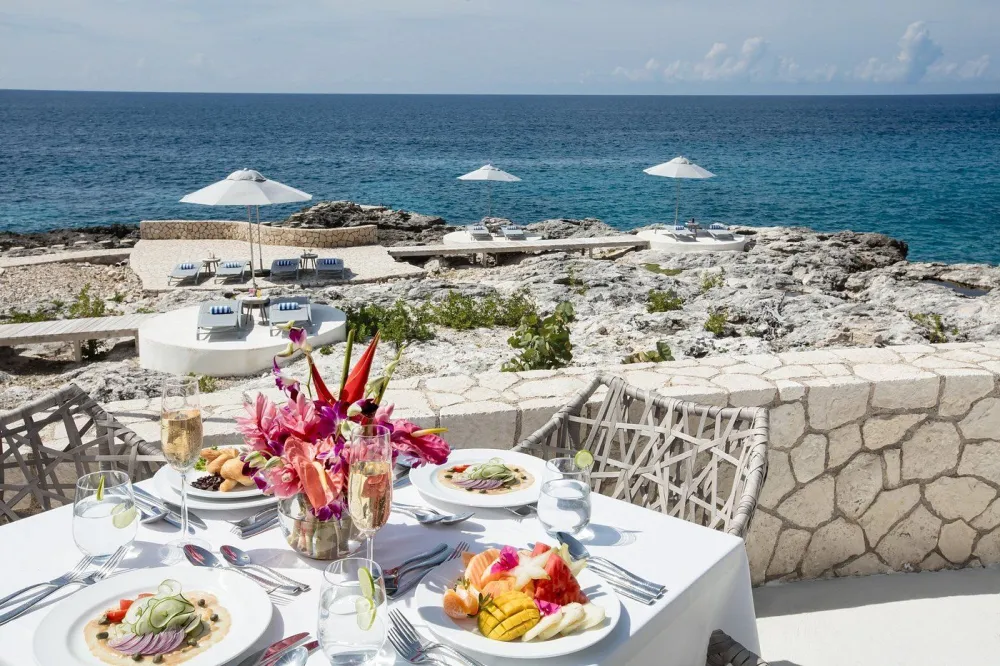 A seaside patio dining scene with a white table set for four, fresh flowers, plates of food, wine glasses, and a stone terrace overlooking the blue ocean under a sunny sky.