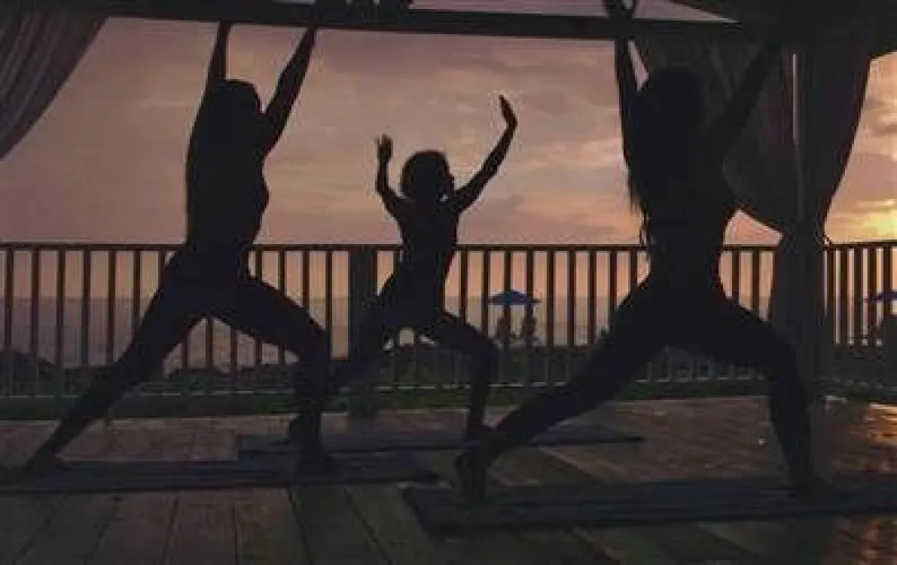 People doing yoga or exercise silhouettes on a wooden deck at sunset, arms raised in celebration, railing and distant horizon in view.