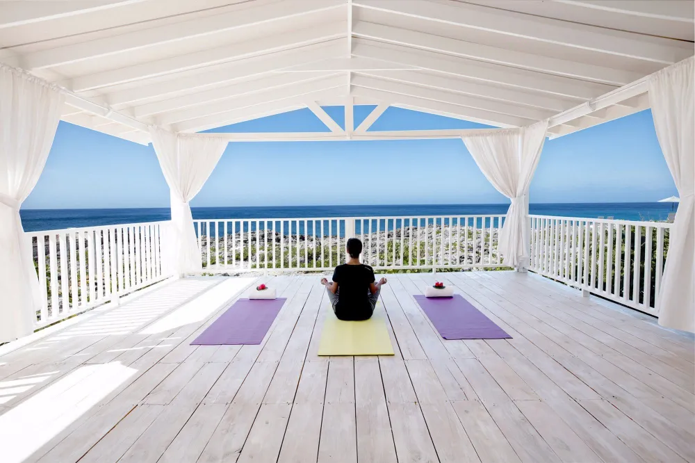 A person sits on a yellow yoga mat under a white gazebo, facing the ocean. Two purple mats flank them on a sunny, open deck.