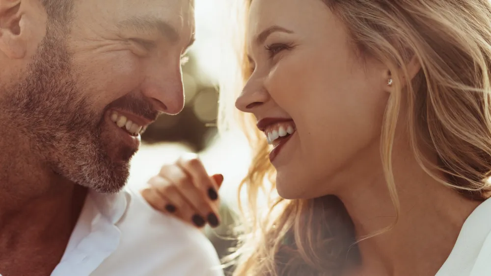 Two adults smiling warmly at each other, close up, suggesting affection or a conversation, with sunlight behind them and a relaxed vibe.