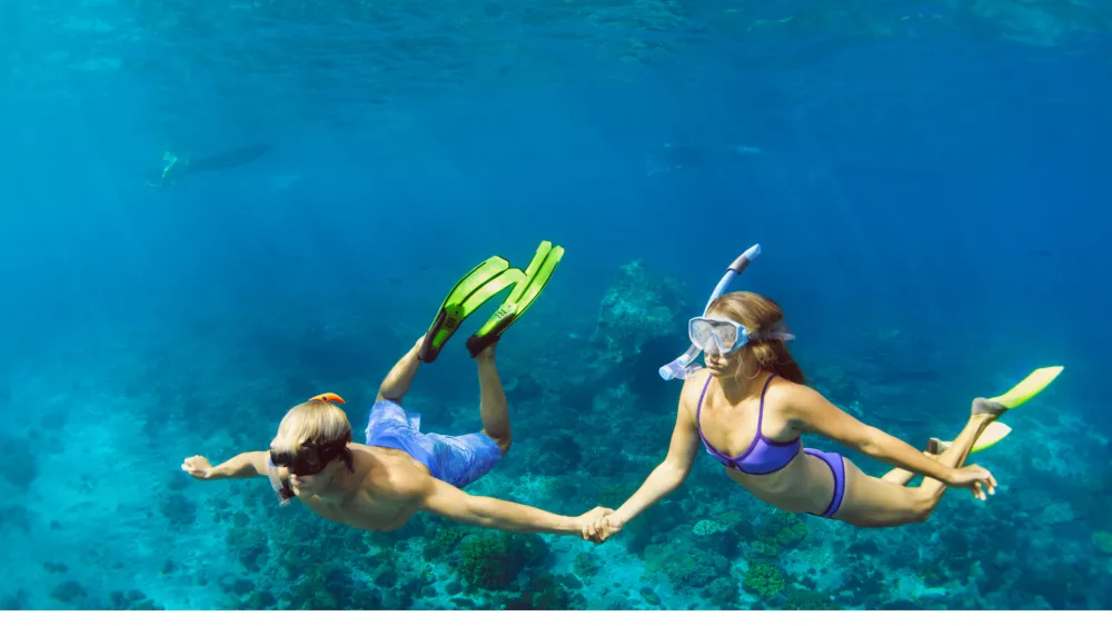 Two people snorkeling underwater, wearing masks and snorkels, holding hands as they swim above a vibrant coral reef with fins. End.