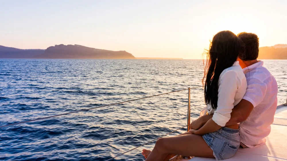 A couple sits close on a boat at sunset, embracing as the calm sea glows golden, with distant landforms and a tranquil horizon. ending the sentence.